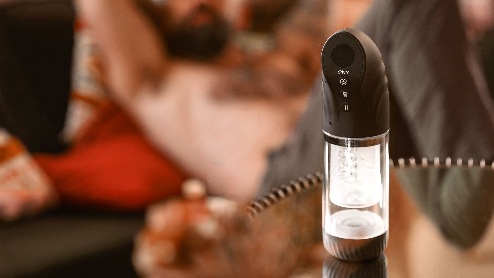 Le masturbateur automatique Exa One posé sur une table. Au second plan, un homme torse-nu couché sur un canapé